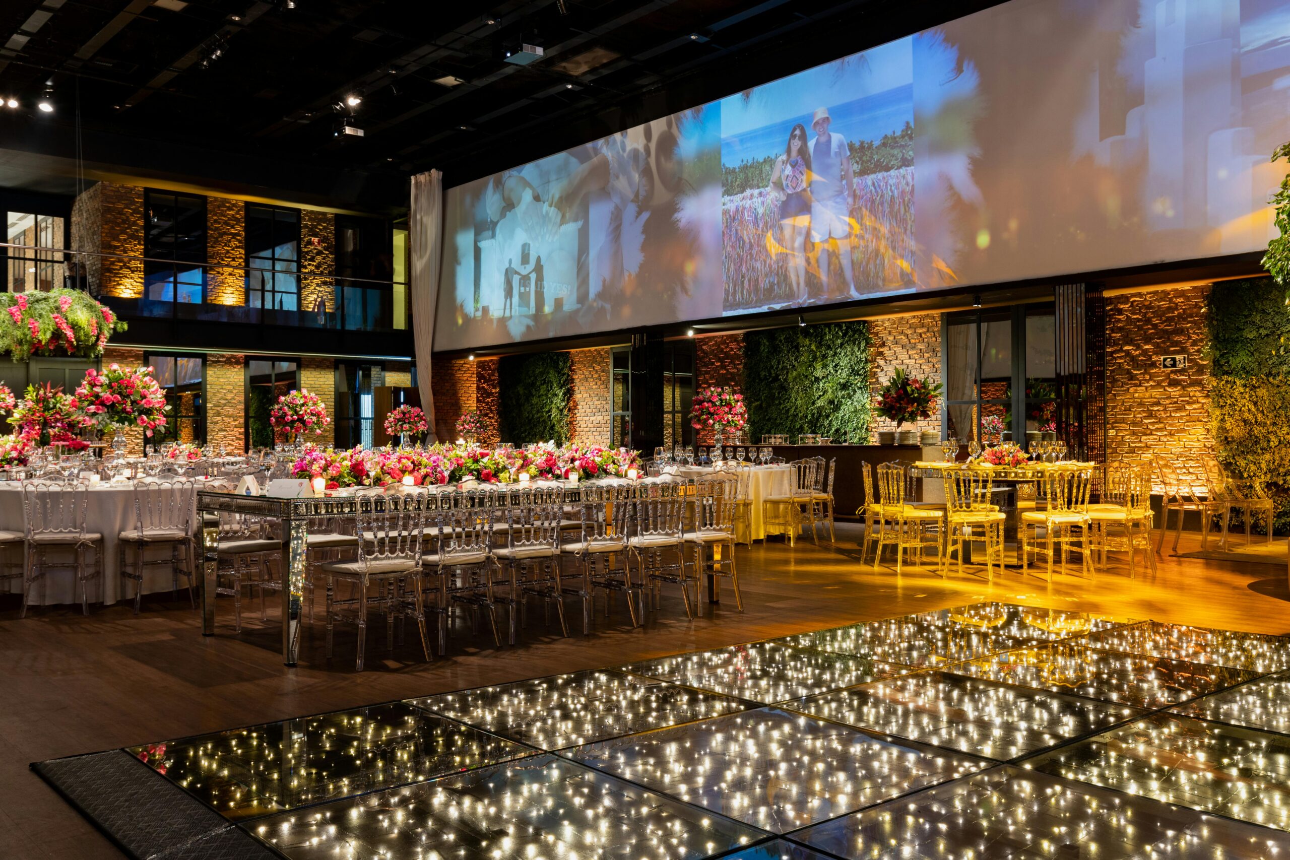 Elegant banquet hall set for a formal event with long tables, pink floral centerpieces, and clear acrylic chairs under warm lighting.